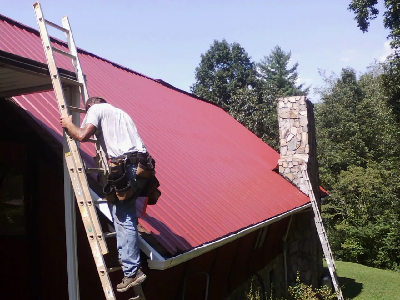 Roofer on a ladder beside a red metal roof and stone chimney — Soffit & Fascia Repair by Roofing Brothers in Amelia Court House